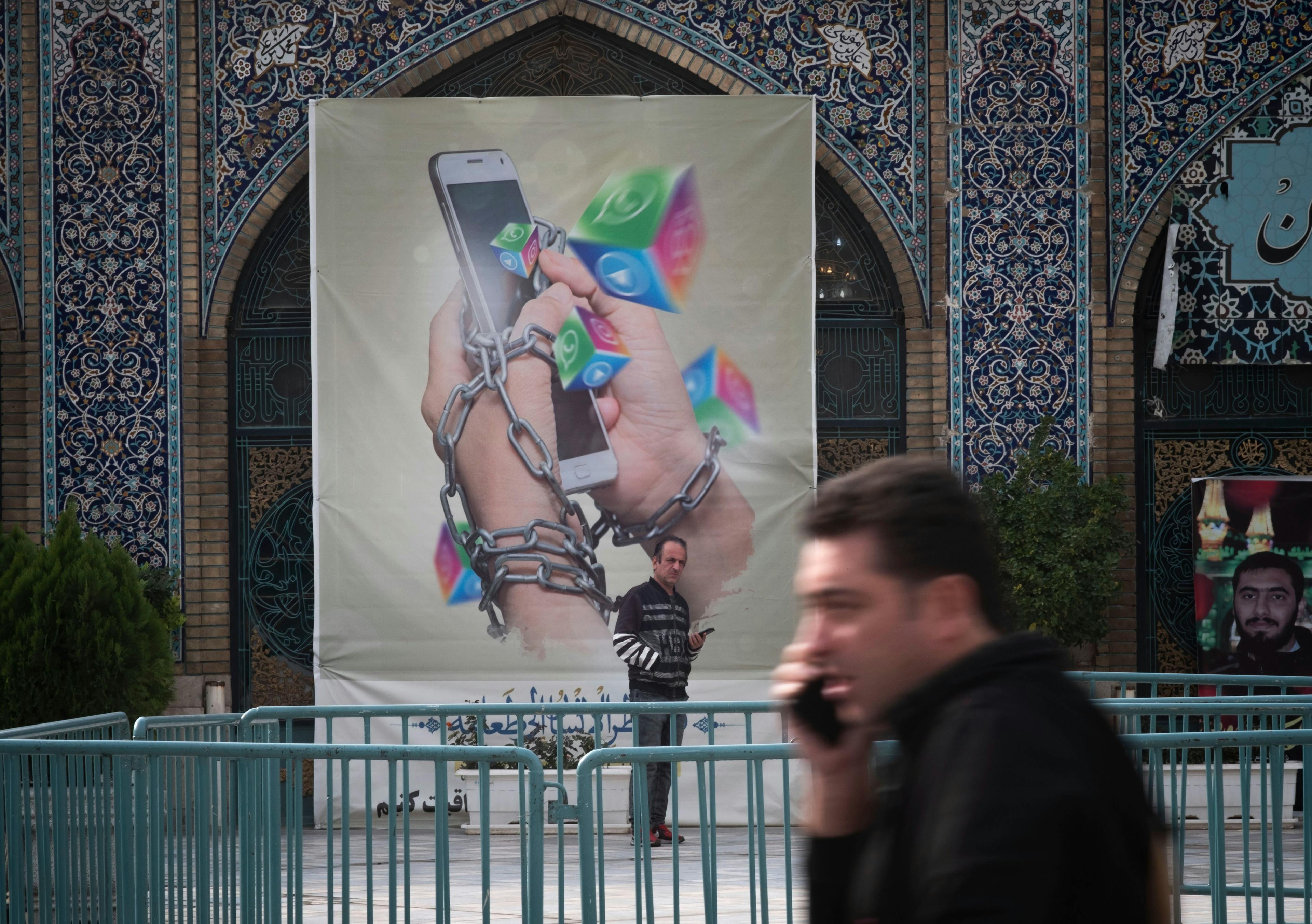 An Iranian man holding his smartphone looks on while standing under an anti-Social networking banner in Tehran Grand Bazaar